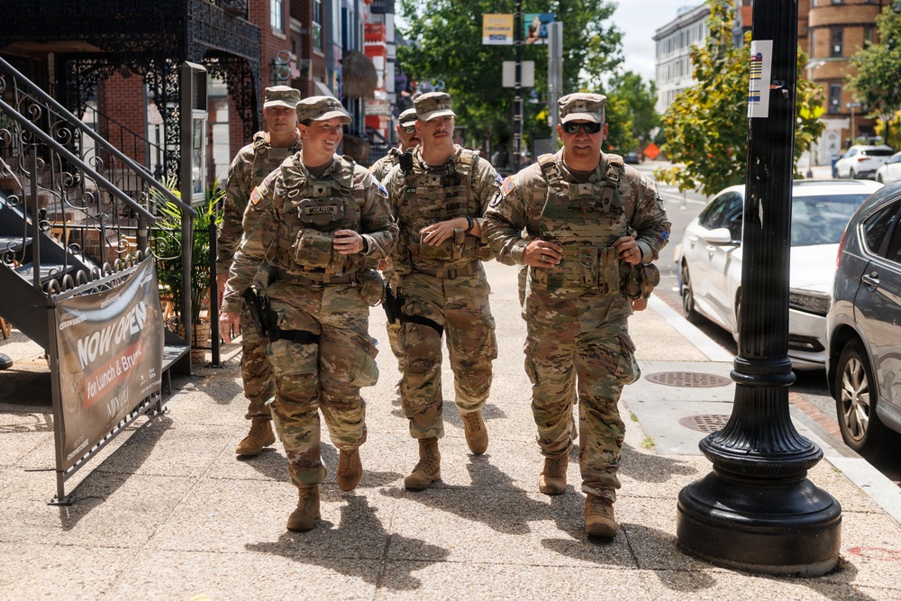 Four U.S. Army soldiers in full camouflage (OCP/MultiCam) uniforms and tactical gear are walking together on a city sidewalk. They appear relaxed and in good spirits, with at least one soldier smiling. The urban setting includes brick buildings, parked cars, and commercial storefronts, suggesting a patrol or presence mission in a metropolitan area.