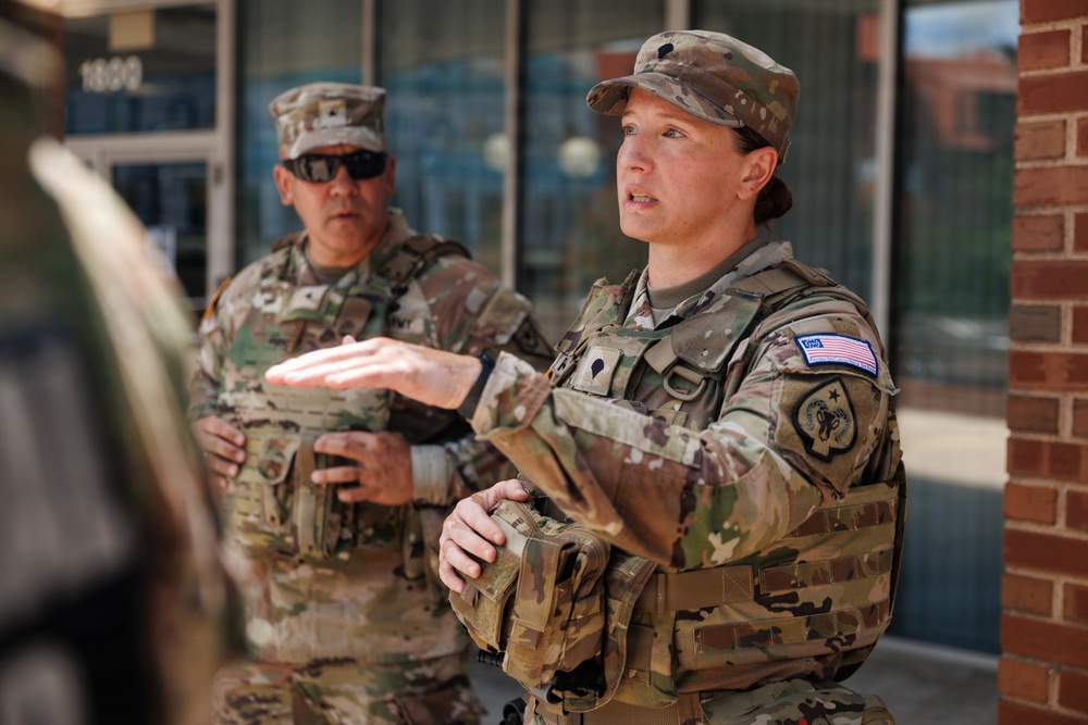 Two U.S. Army soldiers in Operational Camouflage Pattern (OCP) uniforms are standing outside a brick building. The soldier in the foreground, a woman, appears to be speaking and gesturing with her hand, suggesting she is briefing or instructing. Both soldiers are wearing tactical vests and patrol caps.