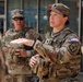 Two U.S. Army soldiers in Operational Camouflage Pattern (OCP) uniforms are standing outside a brick building. The soldier in the foreground, a woman, appears to be speaking and gesturing with her hand, suggesting she is briefing or instructing. Both soldiers are wearing tactical vests and patrol caps.