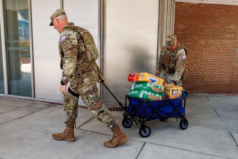 Two military personnel in OCP (Operational Camouflage Pattern) uniforms are transporting supplies on a blue collapsible utility wagon along a sidewalk outside a building. The wagon is loaded with consumer beverages and food items including Fanta, Sprite, and Hug juice boxes. This appears to be a logistics or relief supply distribution activity.