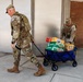 Two military personnel in OCP (Operational Camouflage Pattern) uniforms are transporting supplies on a blue collapsible utility wagon along a sidewalk outside a building. The wagon is loaded with consumer beverages and food items including Fanta, Sprite, and Hug juice boxes. This appears to be a logistics or relief supply distribution activity.