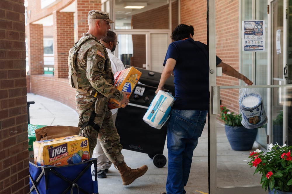 A U.S. Army soldier in camouflage uniform and a civilian in a blue shirt are carrying supplies into a brick building. They are transporting items including paper towels, snack boxes, and a cart loaded with Little Hug Fruit Barrels drinks. A Nexgrill barbecue grill on wheels is also being brought in, suggesting a community event or outreach activity.