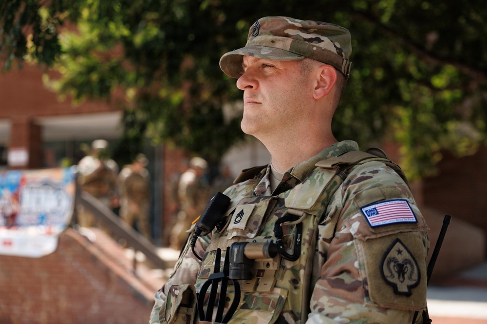 A U.S. Army soldier in full OCP (Operational Camouflage Pattern) uniform stands at attention in an outdoor urban setting. He is wearing a tactical vest with radio equipment and displays rank insignia and unit patches, including a U.S. flag patch on his right shoulder. Additional soldiers are visible in the blurred background, suggesting a formation or security detail.