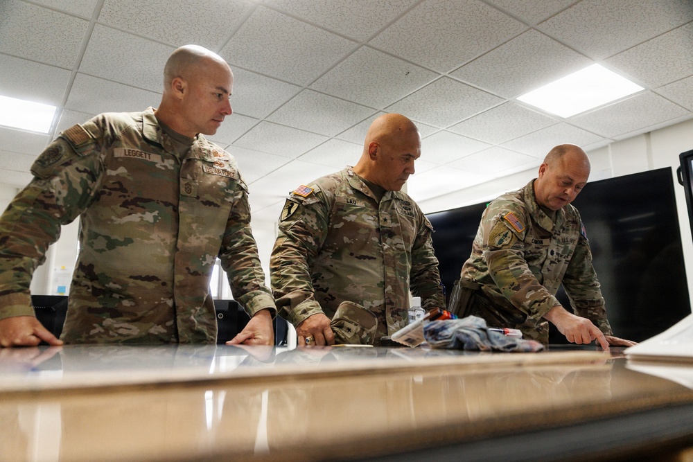Three U.S. military personnel in OCP (Operational Camouflage Pattern) uniforms are gathered around a table, reviewing documents or maps. Name tapes visible on the uniforms read 'LEGGETT', 'LAU', and 'CANON'. The setting appears to be an indoor operations or briefing room with fluorescent lighting.
