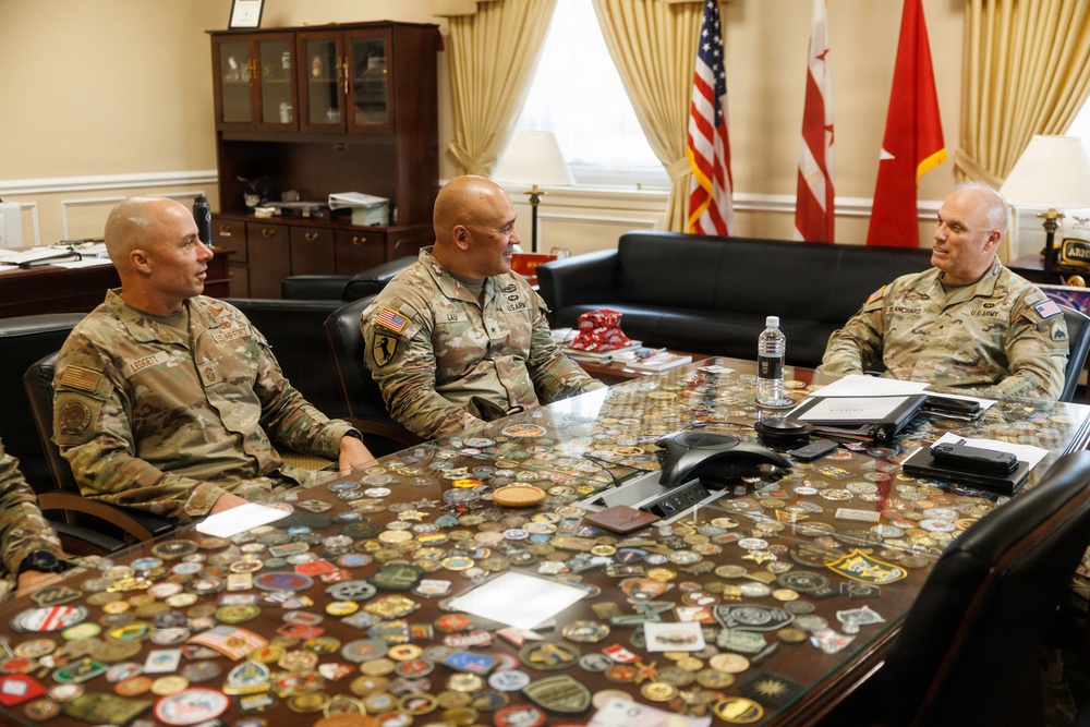 Three U.S. Army soldiers in OCP (Operational Camouflage Pattern) uniforms are seated around a large conference table in a formal military office setting. The glass-topped table displays an extensive collection of military challenge coins and unit patches beneath its surface. The setting appears to be a senior officer's office, indicated by the formal furnishings, multiple flags including the U.S. flag and what appears to be a general officer's flag.