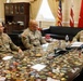 Three U.S. Army soldiers in OCP (Operational Camouflage Pattern) uniforms are seated around a large conference table in a formal military office setting. The glass-topped table displays an extensive collection of military challenge coins and unit patches beneath its surface. The setting appears to be a senior officer's office, indicated by the formal furnishings, multiple flags including the U.S. flag and what appears to be a general officer's flag.