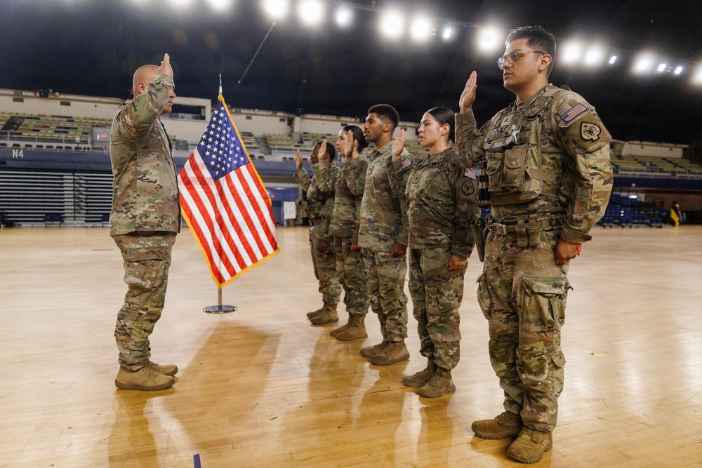 A military re-enlistment or oath of office ceremony is taking place inside what appears to be an indoor arena or gymnasium. A senior officer on the left is administering the oath while four soldiers stand in a line with their right hands raised. An American flag on a stand serves as the ceremonial backdrop.