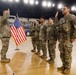 A military re-enlistment or oath of office ceremony is taking place inside what appears to be an indoor arena or gymnasium. A senior officer on the left is administering the oath while four soldiers stand in a line with their right hands raised. An American flag on a stand serves as the ceremonial backdrop.