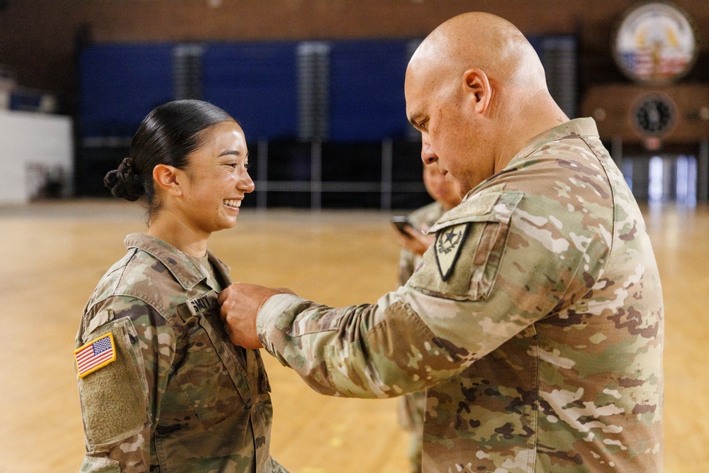 A military ceremony is taking place in what appears to be a gymnasium or indoor facility. A senior soldier is pinning an award or rank insignia onto the uniform of a smiling female soldier. The female soldier displays a visible US Army flag patch on her left shoulder and appears pleased during the ceremony.