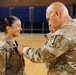 A military ceremony is taking place in what appears to be a gymnasium or indoor facility. A senior soldier is pinning an award or rank insignia onto the uniform of a smiling female soldier. The female soldier displays a visible US Army flag patch on her left shoulder and appears pleased during the ceremony.