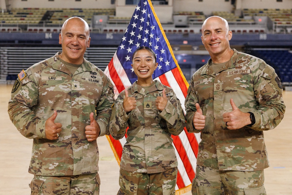 Three U.S. military personnel in OCP (Operational Camouflage Pattern) uniforms pose in front of an American flag inside what appears to be an indoor sports arena. The individuals are giving thumbs-up gestures and smiling. Name tapes and branch insignia are visible on their uniforms, identifying two as U.S. Army and one as U.S. Air Force.