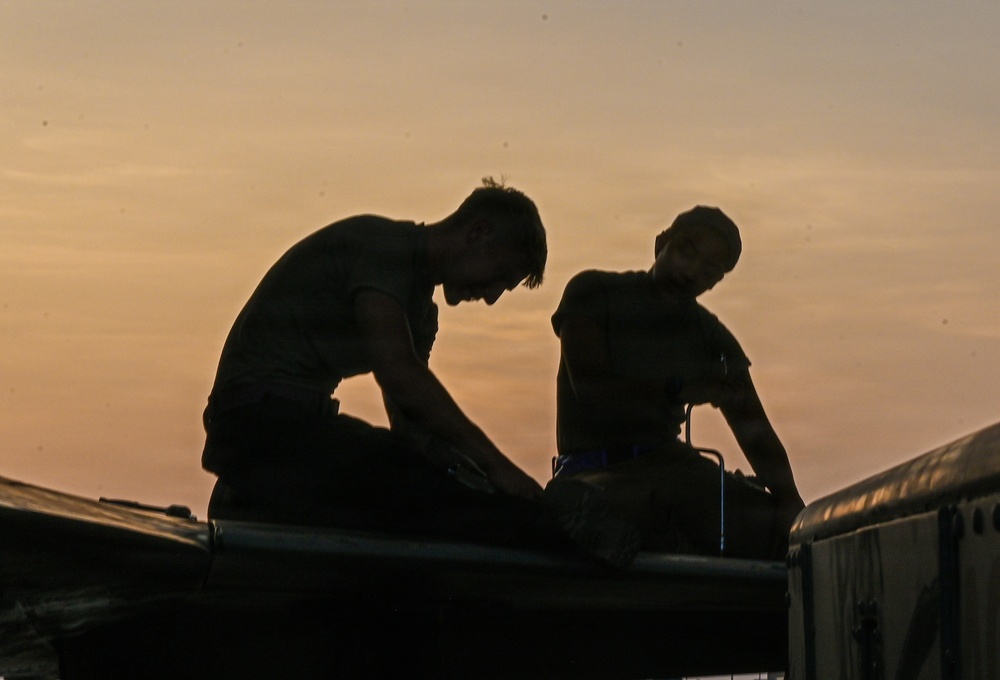 Two individuals are silhouetted against a warm orange and yellow sunset sky. They appear to be working on or handling equipment atop a large vehicle or structure. The strong backlight creates a dramatic silhouette effect, obscuring fine details.