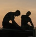 Two individuals are silhouetted against a warm orange and yellow sunset sky. They appear to be working on or handling equipment atop a large vehicle or structure. The strong backlight creates a dramatic silhouette effect, obscuring fine details.