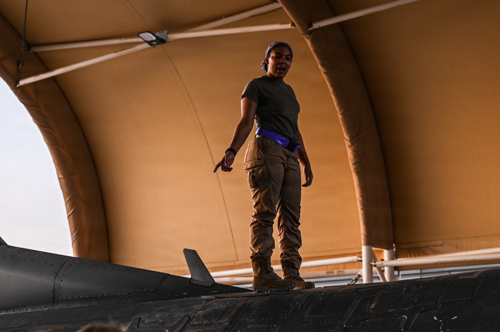 A military service member in olive drab t-shirt and tan cargo pants stands atop a dark-colored military aircraft inside a tan fabric-covered hangar structure. The individual appears to be speaking or giving direction, possibly conducting a pre-flight or maintenance inspection. The setting suggests a forward operating base or deployed location based on the portable hangar construction.