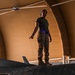 A military service member in olive drab t-shirt and tan cargo pants stands atop a dark-colored military aircraft inside a tan fabric-covered hangar structure. The individual appears to be speaking or giving direction, possibly conducting a pre-flight or maintenance inspection. The setting suggests a forward operating base or deployed location based on the portable hangar construction.