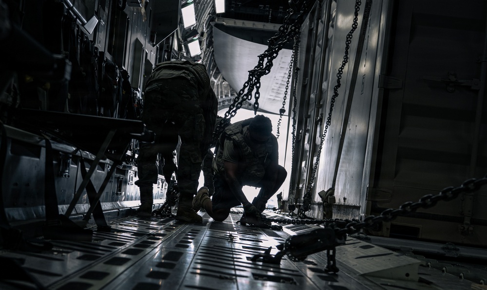 The image shows the interior of a military cargo aircraft, likely a C-130 or C-17, with two personnel in military uniforms and tactical gear working inside the cargo hold. Heavy chains used for securing cargo are visible running along the sides and floor of the aircraft. The scene is dramatically lit with strong contrast between shadow and ambient light entering from the open rear ramp.