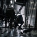 The image shows the interior of a military cargo aircraft, likely a C-130 or C-17, with two personnel in military uniforms and tactical gear working inside the cargo hold. Heavy chains used for securing cargo are visible running along the sides and floor of the aircraft. The scene is dramatically lit with strong contrast between shadow and ambient light entering from the open rear ramp.