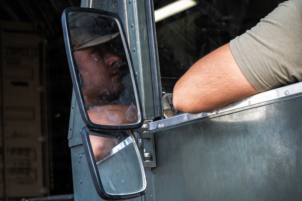 A person wearing a military-style cap is visible in the side mirror of a large military or tactical vehicle. The image is taken from outside the vehicle, showing the driver's reflection in the dual side mirrors. The background suggests a cargo or transport environment with boxes visible.