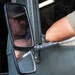 A person wearing a military-style cap is visible in the side mirror of a large military or tactical vehicle. The image is taken from outside the vehicle, showing the driver's reflection in the dual side mirrors. The background suggests a cargo or transport environment with boxes visible.