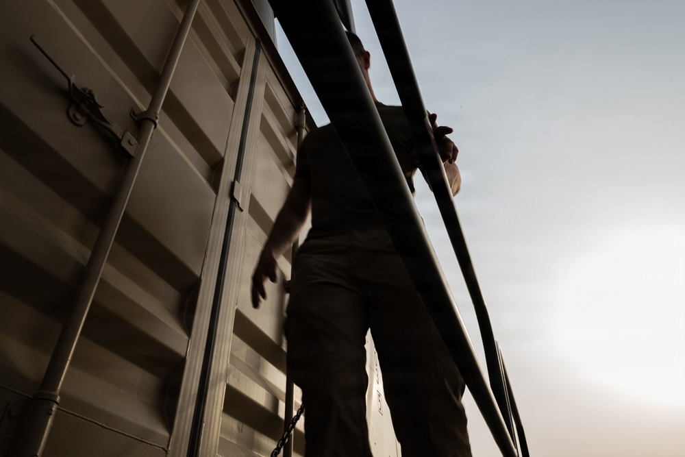 A person is photographed from a low angle, standing alongside what appears to be a large metal freight container or railcar. The image is taken from below, creating a dramatic upward perspective with the figure silhouetted against a hazy sky. The scene suggests the individual is climbing or moving along the side of the structure.