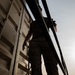 A person is photographed from a low angle, standing alongside what appears to be a large metal freight container or railcar. The image is taken from below, creating a dramatic upward perspective with the figure silhouetted against a hazy sky. The scene suggests the individual is climbing or moving along the side of the structure.