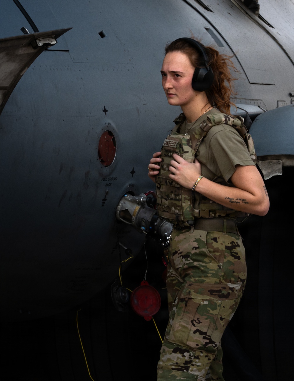 A female military service member in MultiCam/OCP camouflage uniform and plate carrier vest stands beside a gray military aircraft, identifiable by visible 'F-40' and 'FUEL GROUND JACK' stenciling on the fuselage. She is wearing over-ear hearing protection and holding a fuel nozzle/hose assembly, suggesting she is performing aircraft ground servicing duties. The image appears to be a professional or documentary-style portrait taken on a flight line.