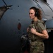 A female military service member in MultiCam/OCP camouflage uniform and plate carrier vest stands beside a gray military aircraft, identifiable by visible 'F-40' and 'FUEL GROUND JACK' stenciling on the fuselage. She is wearing over-ear hearing protection and holding a fuel nozzle/hose assembly, suggesting she is performing aircraft ground servicing duties. The image appears to be a professional or documentary-style portrait taken on a flight line.