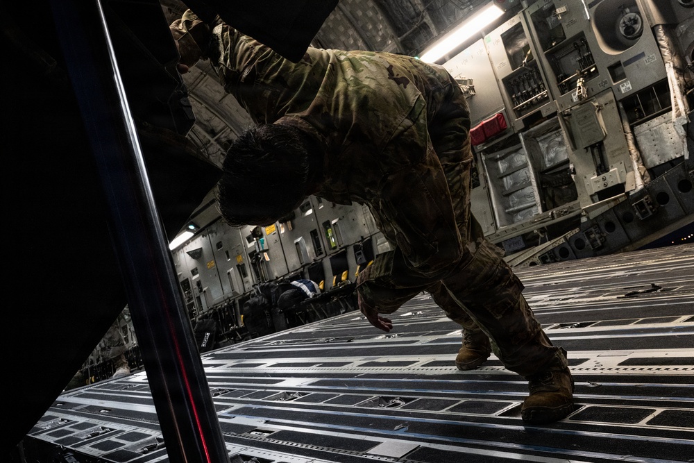 A military service member in camouflage uniform is working inside the cargo hold of a large military transport aircraft, likely a C-17 Globemaster III. The individual appears to be pushing or securing cargo near the aircraft's ramp area. The image is taken from a dramatic low angle, emphasizing the scale of the aircraft interior.