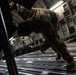 A military service member in camouflage uniform is working inside the cargo hold of a large military transport aircraft, likely a C-17 Globemaster III. The individual appears to be pushing or securing cargo near the aircraft's ramp area. The image is taken from a dramatic low angle, emphasizing the scale of the aircraft interior.