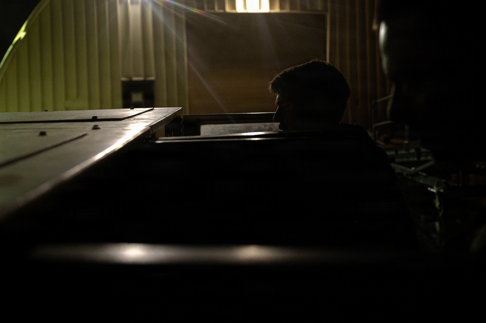 A dimly lit industrial or warehouse setting showing a person in silhouette positioned behind large metal equipment or machinery. The scene is dramatically backlit with a bright light source visible in the background, creating strong contrast between shadow and light. The environment appears to be inside a metal-walled structure, possibly a shipping container or industrial facility.