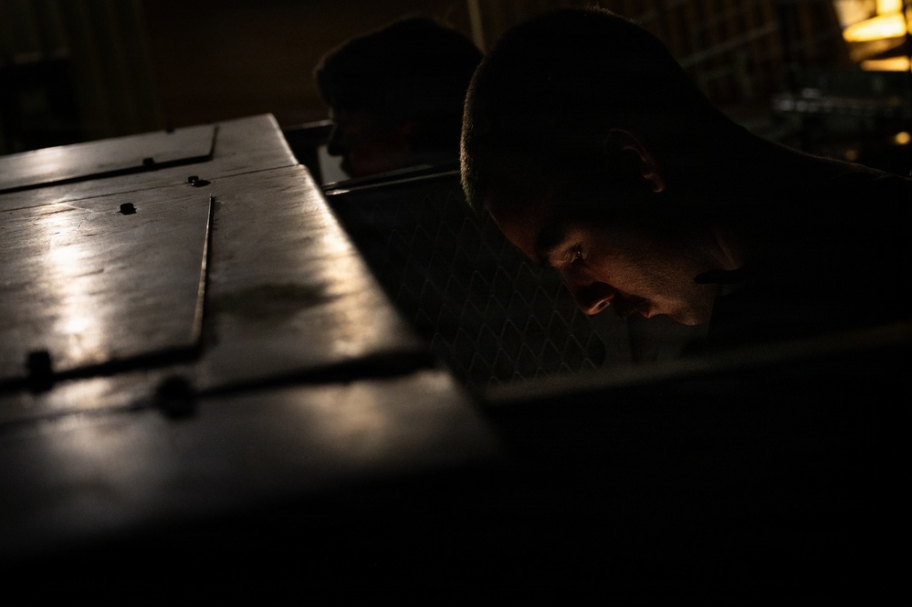 A dramatically lit, low-angle photograph shows a person closely inspecting or examining a dark metal surface or equipment in a dimly lit environment. The image uses strong chiaroscuro lighting, with a single light source illuminating portions of the metal surface and the subject's face. The overall mood is tense and focused, suggesting a detailed inspection or maintenance task.