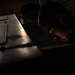 A dramatically lit, low-angle photograph shows a person closely inspecting or examining a dark metal surface or equipment in a dimly lit environment. The image uses strong chiaroscuro lighting, with a single light source illuminating portions of the metal surface and the subject's face. The overall mood is tense and focused, suggesting a detailed inspection or maintenance task.