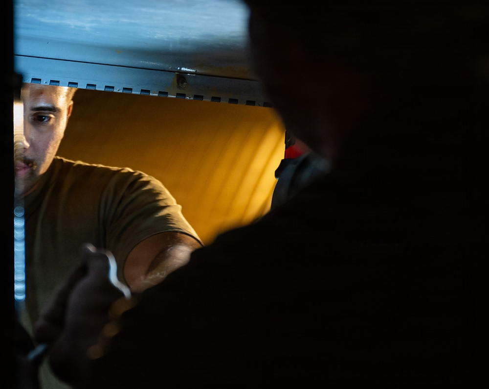 A man wearing an olive/tan colored t-shirt is visible through a narrow opening, likely a hatch or access panel, illuminated by warm amber-yellow light. He appears to be reaching or working with a tool, possibly a wrench or similar implement. The foreground is dominated by a dark silhouette, creating a dramatic contrast with the lit background.
