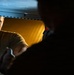 A man wearing an olive/tan colored t-shirt is visible through a narrow opening, likely a hatch or access panel, illuminated by warm amber-yellow light. He appears to be reaching or working with a tool, possibly a wrench or similar implement. The foreground is dominated by a dark silhouette, creating a dramatic contrast with the lit background.