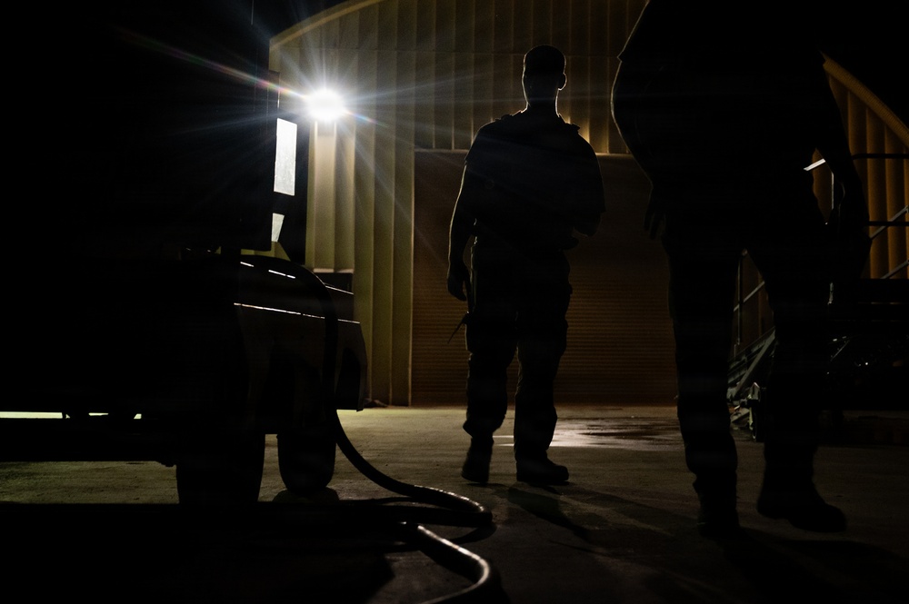 A nighttime scene showing two silhouetted figures standing near an industrial or military building. A bright light source illuminates the background, creating strong contrast against the dark foreground. A hose or cable is visible on the ground near what appears to be equipment or a vehicle.