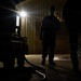 A nighttime scene showing two silhouetted figures standing near an industrial or military building. A bright light source illuminates the background, creating strong contrast against the dark foreground. A hose or cable is visible on the ground near what appears to be equipment or a vehicle.