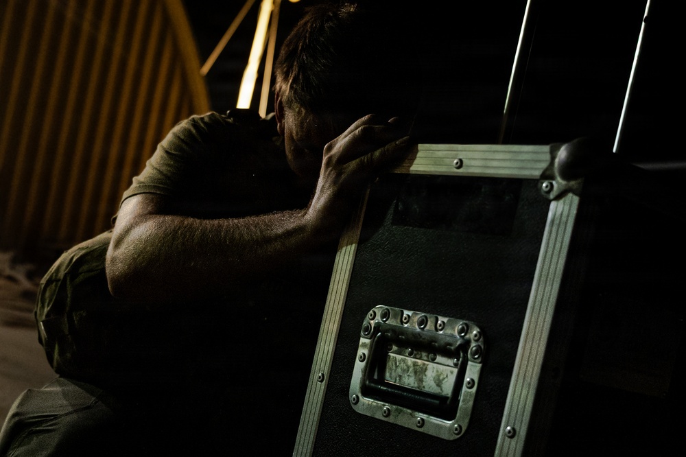 A man in a dark olive/green t-shirt is crouched or leaning against a large black road case or equipment flight case with metal hardware and a recessed handle. The scene appears to be backstage or in a dimly lit venue or warehouse setting. The lighting is dramatic and low-key, with warm tones visible in the background.