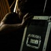 A man in a dark olive/green t-shirt is crouched or leaning against a large black road case or equipment flight case with metal hardware and a recessed handle. The scene appears to be backstage or in a dimly lit venue or warehouse setting. The lighting is dramatic and low-key, with warm tones visible in the background.