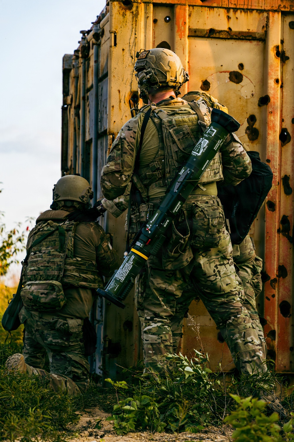 Two soldiers in full tactical gear are taking cover beside a heavily rusted and bullet-riddled metal container or vehicle. The standing soldier carries what appears to be an AT4 or similar single-use anti-armor rocket launcher slung across his back. The scene suggests a tactical movement or clearing operation in a combat or training environment.