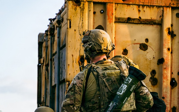 Two soldiers in full tactical gear are taking cover beside a heavily rusted and bullet-riddled metal container or vehicle. The standing soldier carries what appears to be an AT4 or similar single-use anti-armor rocket launcher slung across his back. The scene suggests a tactical movement or clearing operation in a combat or training environment.