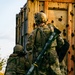 Two soldiers in full tactical gear are taking cover beside a heavily rusted and bullet-riddled metal container or vehicle. The standing soldier carries what appears to be an AT4 or similar single-use anti-armor rocket launcher slung across his back. The scene suggests a tactical movement or clearing operation in a combat or training environment.