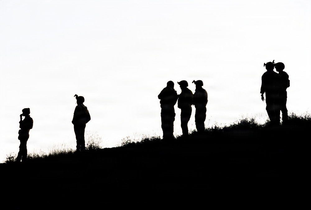 The image shows silhouettes of approximately seven individuals positioned along the ridge of a hill against a bright, overcast sky. The figures appear to be equipped with gear including helmets and backpacks, suggesting a military or tactical context. The high-contrast backlighting renders all subjects as complete black silhouettes with no distinguishing color or facial detail visible.