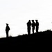 The image shows silhouettes of approximately seven individuals positioned along the ridge of a hill against a bright, overcast sky. The figures appear to be equipped with gear including helmets and backpacks, suggesting a military or tactical context. The high-contrast backlighting renders all subjects as complete black silhouettes with no distinguishing color or facial detail visible.