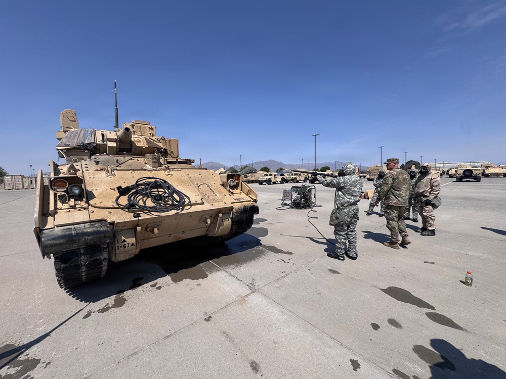 Military personnel in camouflage uniforms stand near a Bradley Fighting Vehicle (IFV) on a paved motor pool area at what appears to be a U.S. Army installation in a desert environment. The vehicle bears the marking '1-67' and shows signs of field use with dust and wear. Several soldiers appear to be conducting maintenance or inspection activities with equipment connected by hoses.