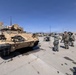 Military personnel in camouflage uniforms stand near a Bradley Fighting Vehicle (IFV) on a paved motor pool area at what appears to be a U.S. Army installation in a desert environment. The vehicle bears the marking '1-67' and shows signs of field use with dust and wear. Several soldiers appear to be conducting maintenance or inspection activities with equipment connected by hoses.
