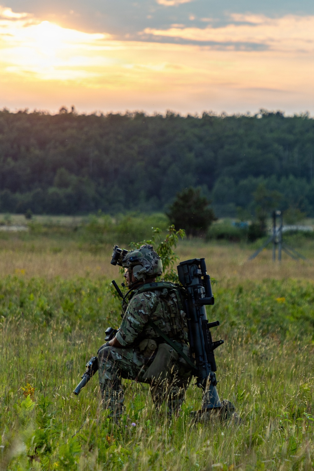 A soldier in full combat gear is positioned in a grassy field at sunset or sunrise, facing away from the camera toward a treeline. The soldier is equipped with a helmet, camouflage uniform, tactical vest, and what appears to be a man-portable air-defense system (MANPADS) or similar shoulder-launched weapon system mounted on their back. The setting appears to be a training exercise or operational deployment in a temperate environment.
