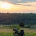A soldier in full combat gear is positioned in a grassy field at sunset or sunrise, facing away from the camera toward a treeline. The soldier is equipped with a helmet, camouflage uniform, tactical vest, and what appears to be a man-portable air-defense system (MANPADS) or similar shoulder-launched weapon system mounted on their back. The setting appears to be a training exercise or operational deployment in a temperate environment.