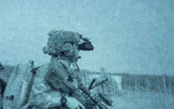The image shows two heavily equipped soldiers in full combat gear standing in an open field with tall grass. The photo has a distinctive blue-tinted, grainy quality consistent with night-vision device photography or a thermal/NVG-filtered camera. The soldiers appear to be on patrol or conducting a tactical movement.