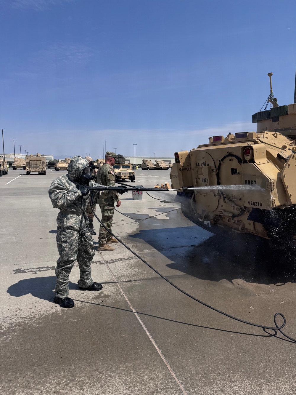 A U.S. Army soldier wearing full MOPP (Mission Oriented Protective Posture) gear and a gas mask is pressure-washing a Bradley Fighting Vehicle in a military motor pool. A second soldier in standard ACU/OCP uniform stands nearby observing or supervising. Multiple military vehicles are visible in the background of what appears to be a large paved staging area.