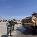 A U.S. Army soldier wearing full MOPP (Mission Oriented Protective Posture) gear and a gas mask is pressure-washing a Bradley Fighting Vehicle in a military motor pool. A second soldier in standard ACU/OCP uniform stands nearby observing or supervising. Multiple military vehicles are visible in the background of what appears to be a large paved staging area.
